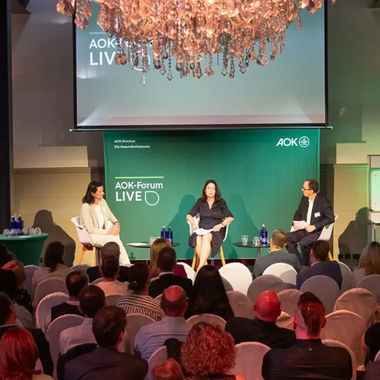 AOK Forum live mit Britta Müller, Daniela Teichert, moderiert von Marcel Weigand Podiumsdiskussion mit drei Teilnehmenden. Britta Müller mittig, Daniela Teichert links und Marcel Weigand rechts auf der Bühne.
